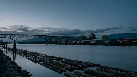 A wide river at dusk with a log boom along the bank, a steel truss bridge and low buildings with lit windows on the far shore.